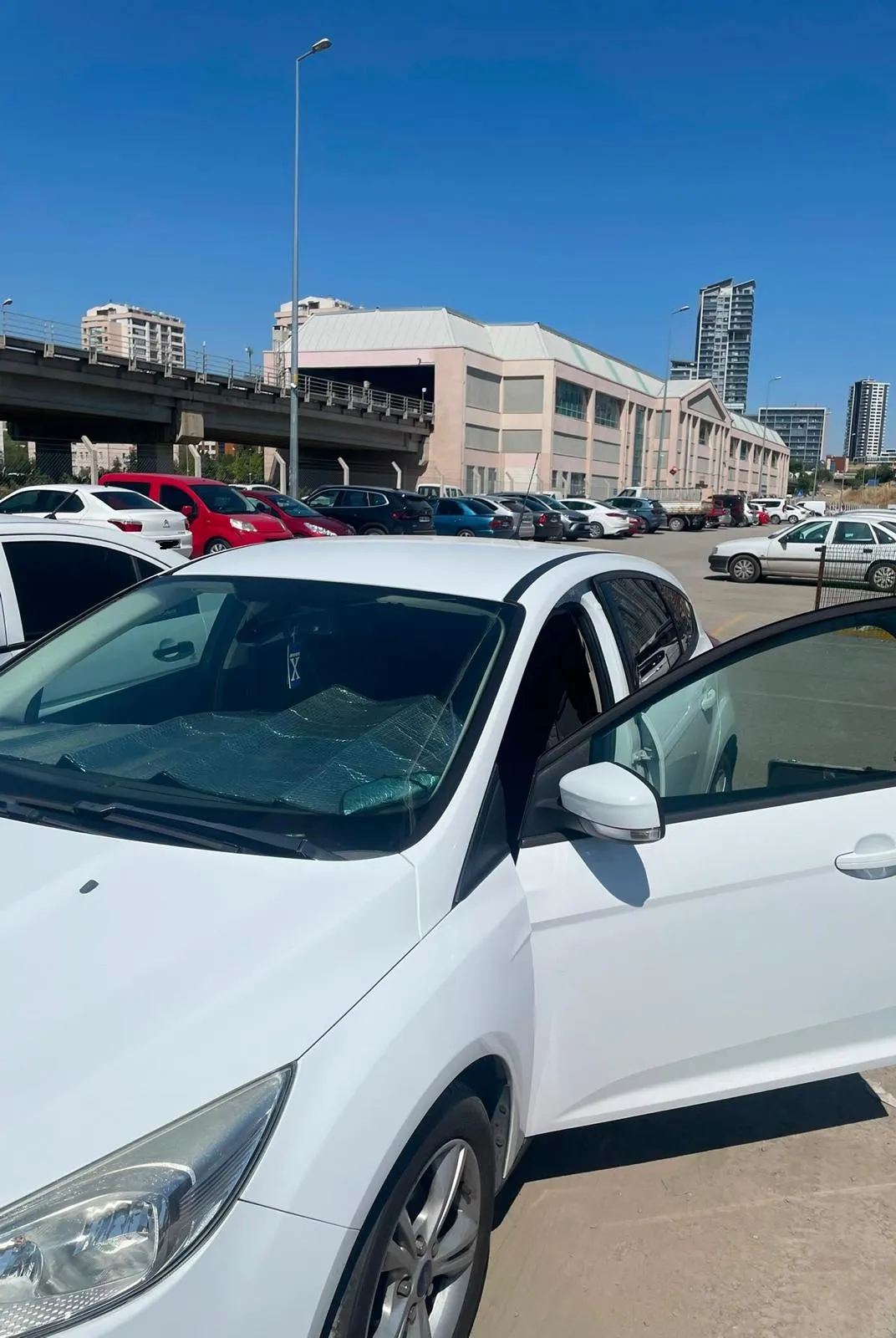 Geniş açık otoparkta kapısı açılmış beyaz Ford Focus; arkada viyadük ve alışveriş merkezi