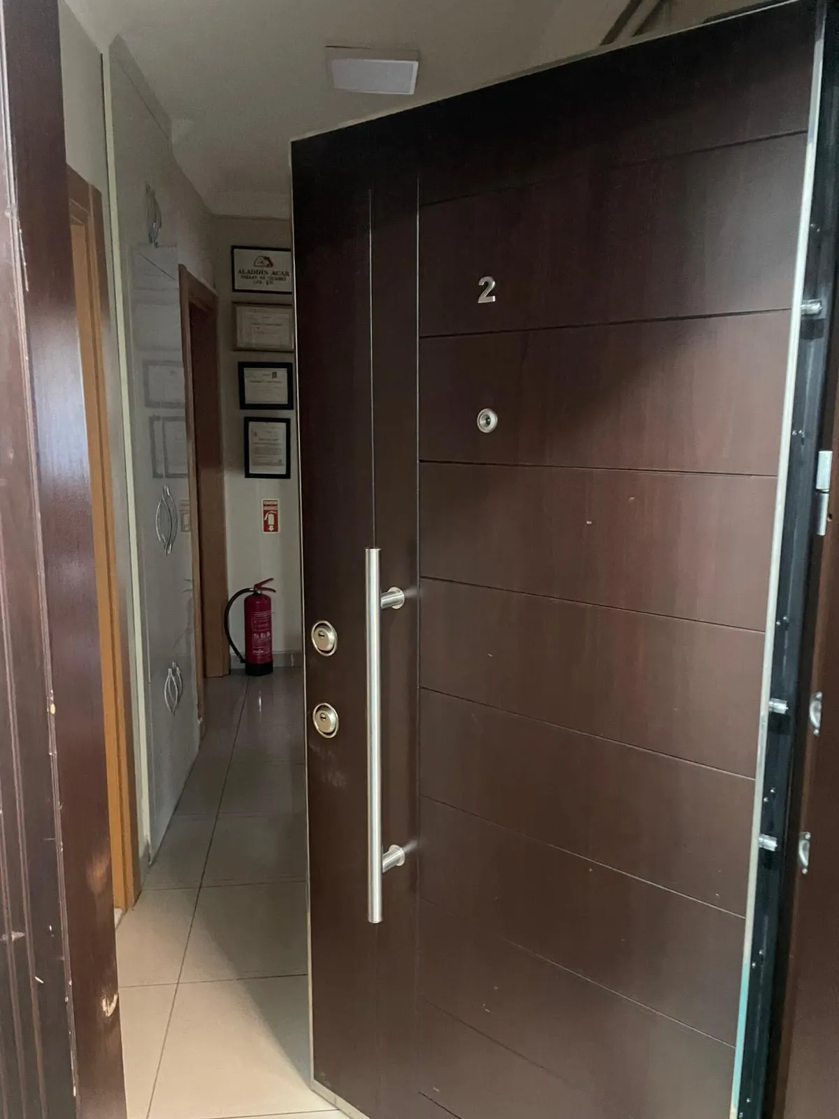 Apartman koridorunda koyu kahverengi çelik daire kapısı; iki barel rozeti, boru kulp ve gözetleme deliği