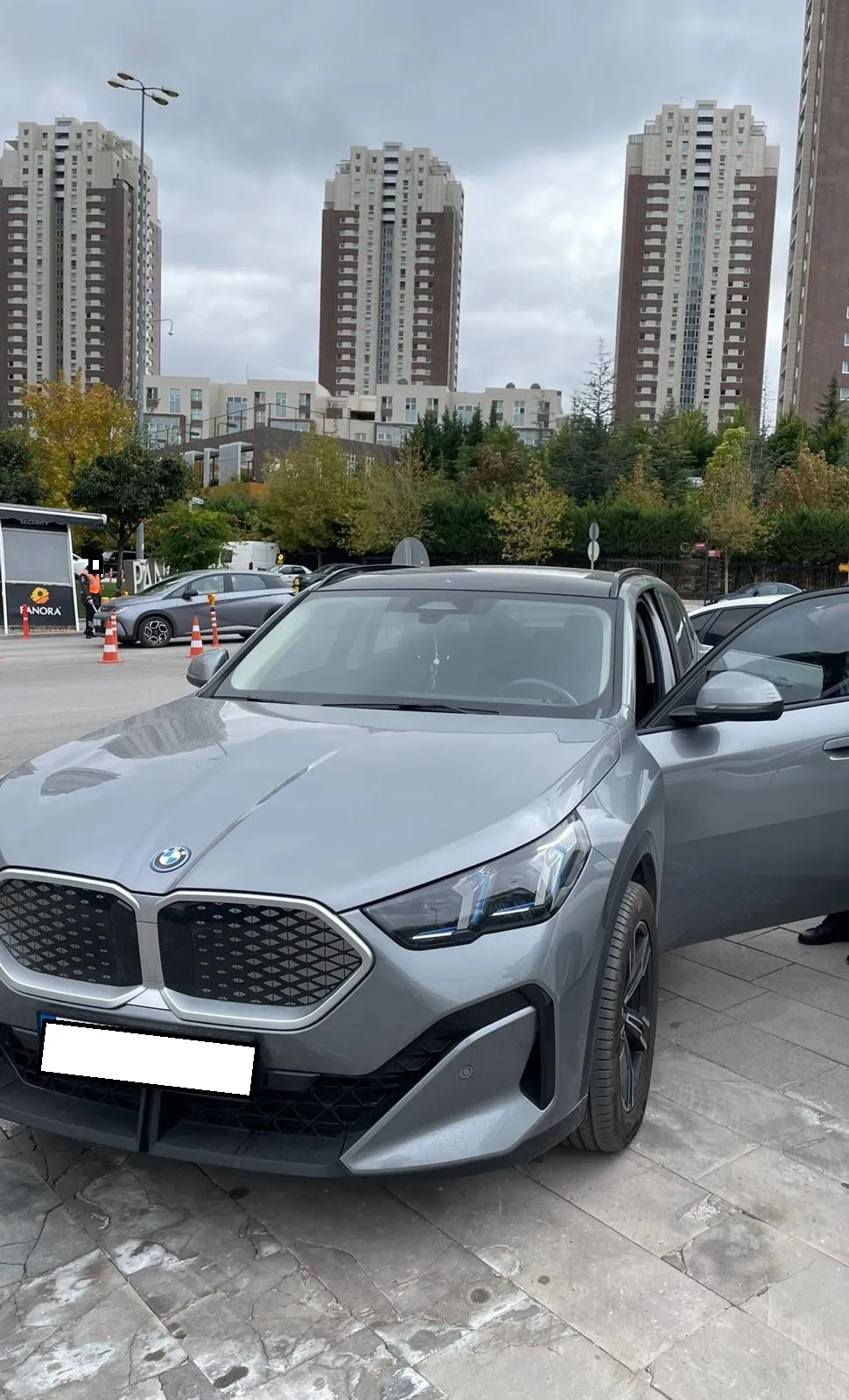 AVM otoparkında kapısı açılmış gri BMW; arkada alışveriş merkezi binası ve otopark görevlileri
