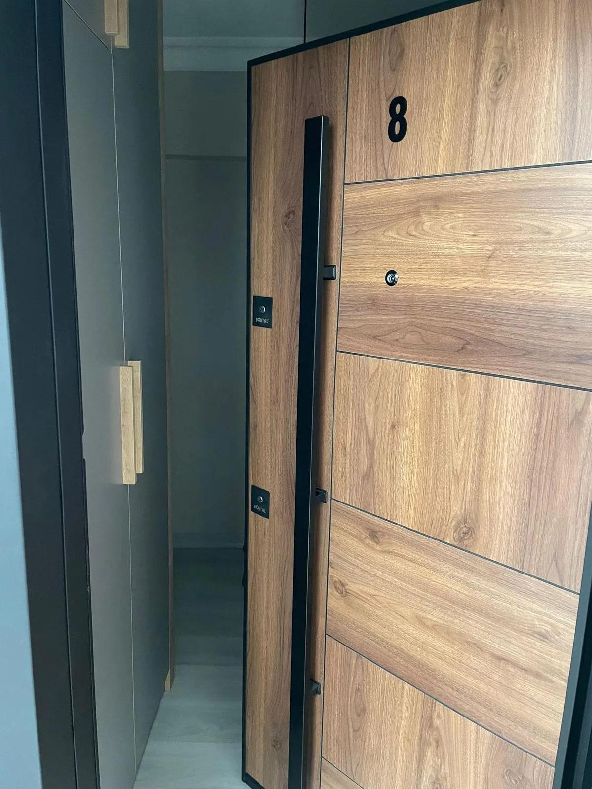 Montajı tamamlanmış ahşap desenli çelik daire kapısı: siyah kasa, uzun barel kulp, gözetleme deliği ve daire numarası