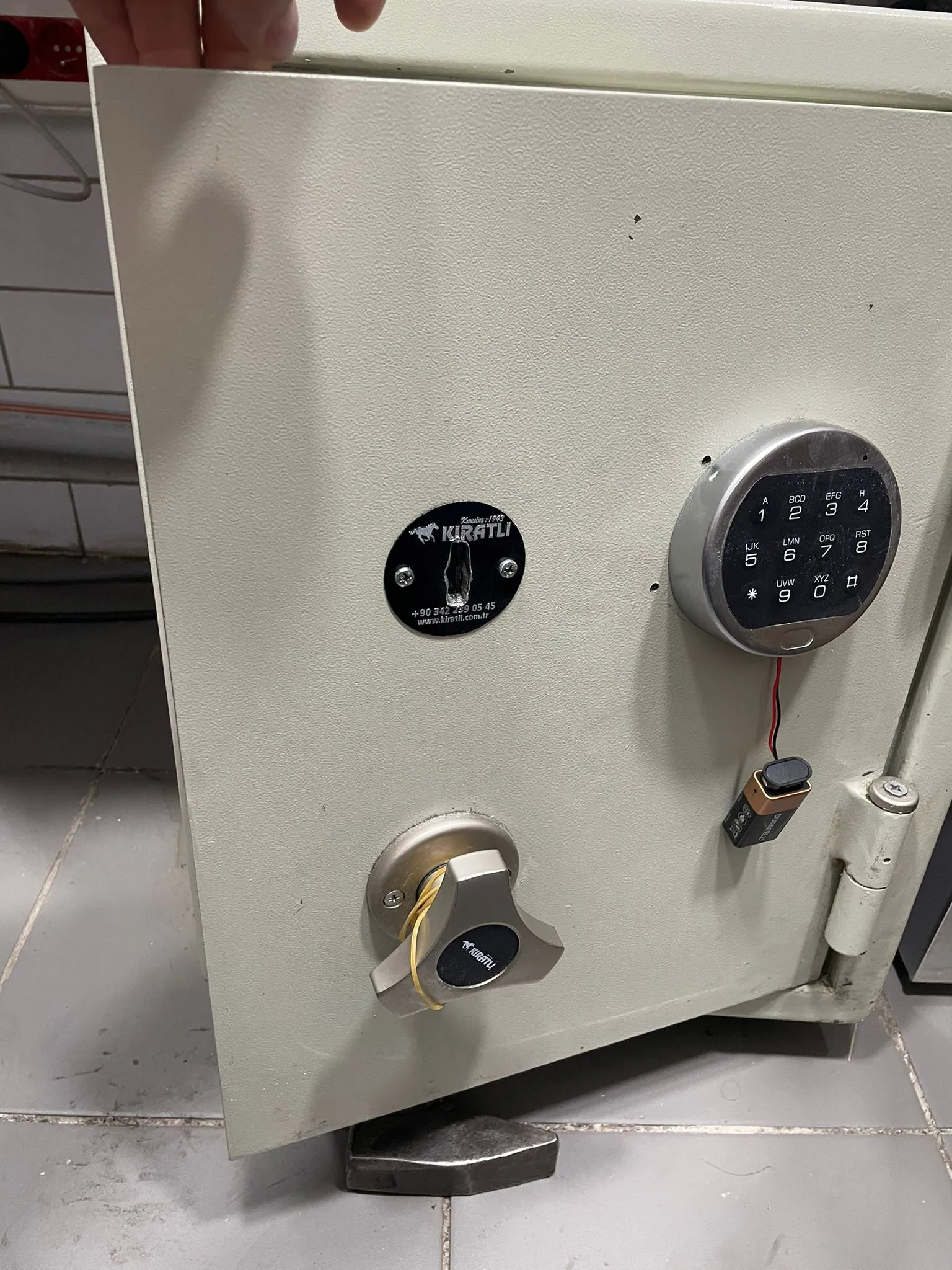Kıratlı marka çelik kasanın kapağında elektronik şifre tuş takımı; dışarıdan bağlanmış 9 voltluk pil ve krom kol görünüyor