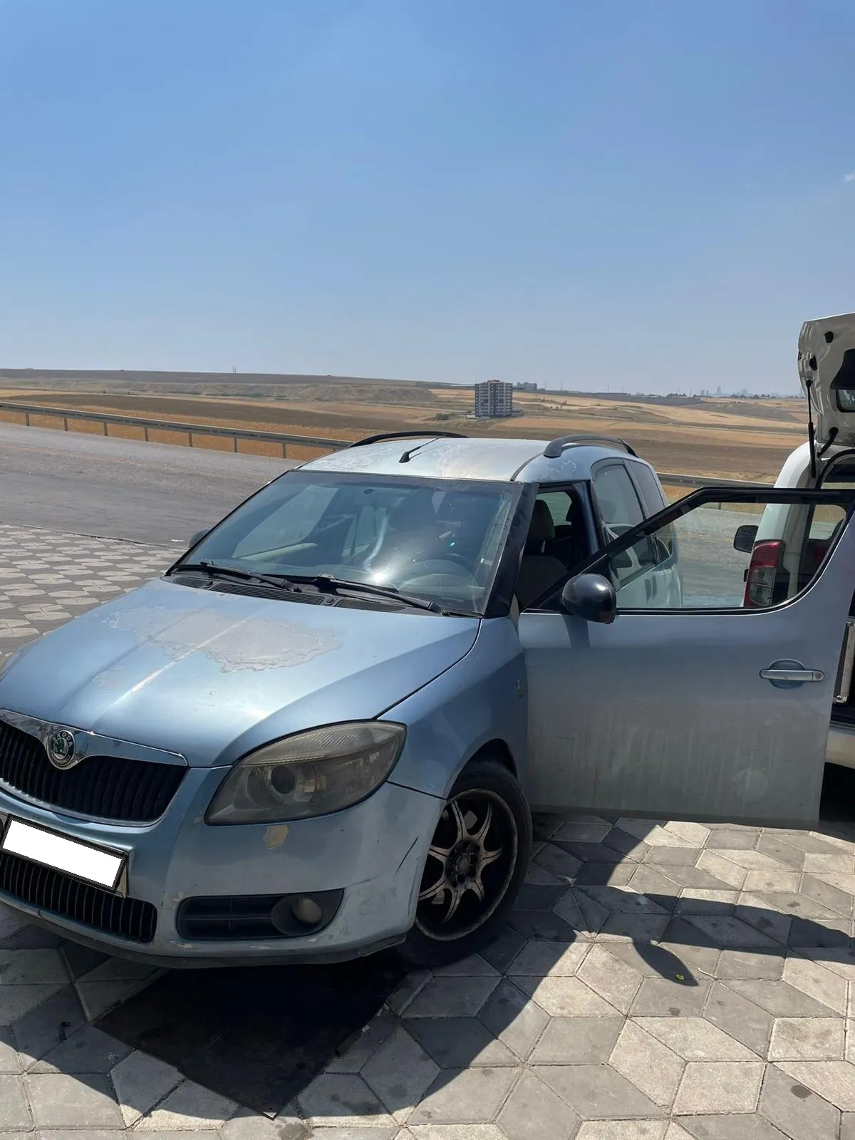 Şehirlerarası yol kenarında kapısı açılmış açık mavi Škoda ve arkasında bagajı açık servis aracı